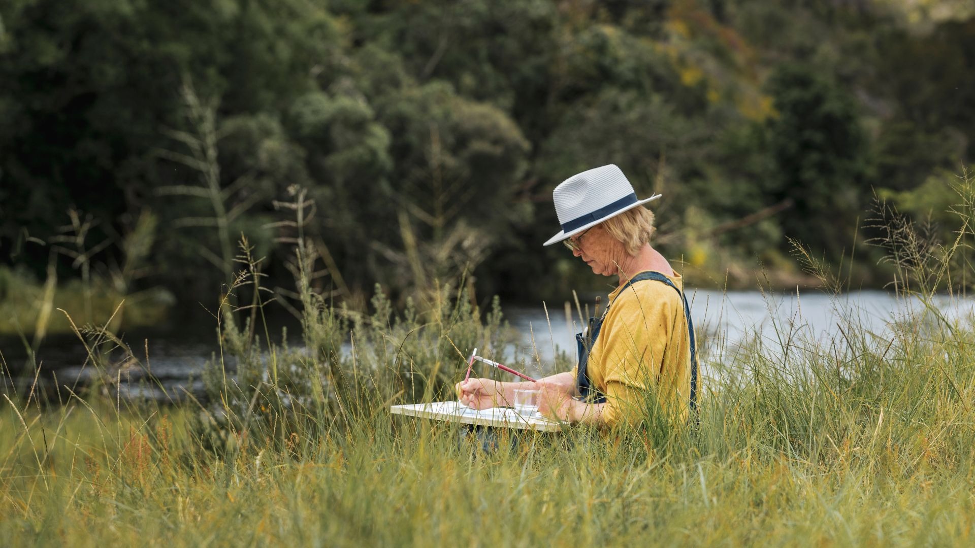 A woman artist wearing a hat paints on a canvas while sitting in a grassy field by a river.