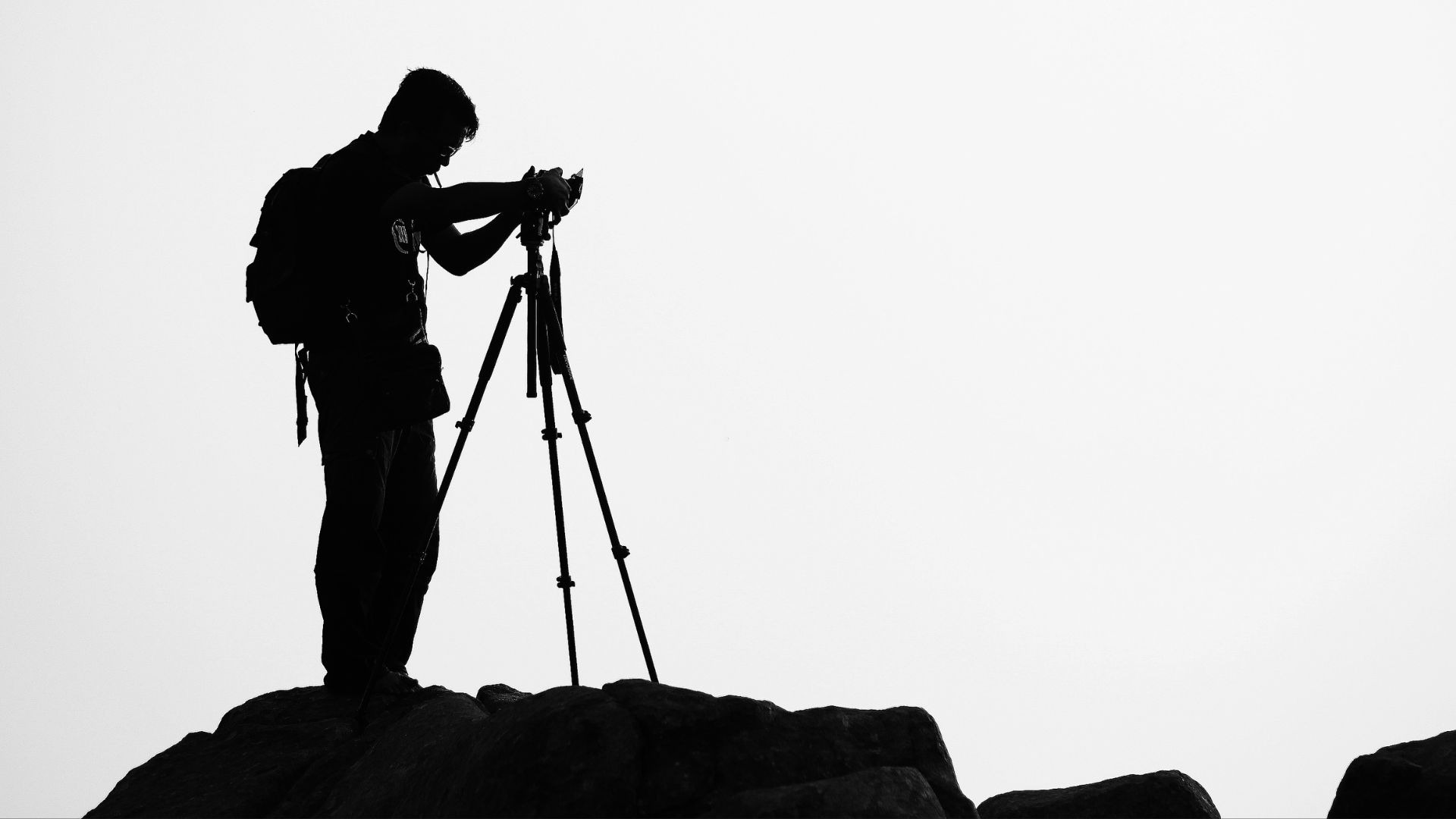 Silhouette of a travel photographer with a backpack using a tripod and camera on a rocky peak.